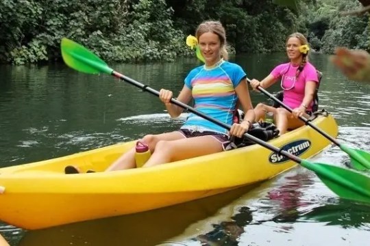 Two women kayaking on a river, wearing colorful shirts and holding paddles, surrounded by lush greenery.
