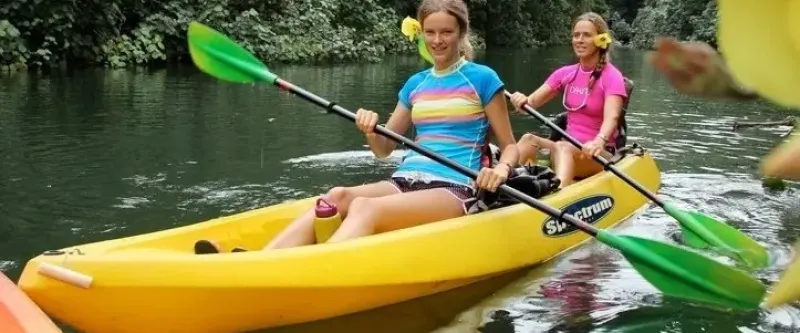 Two women kayaking on a river, wearing colorful shirts and holding paddles, surrounded by lush greenery.