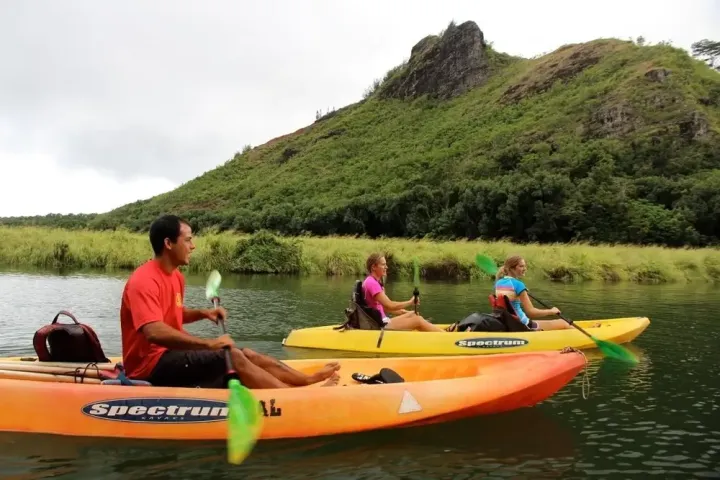 Three people kayaking on a calm river near a green, rocky hill.